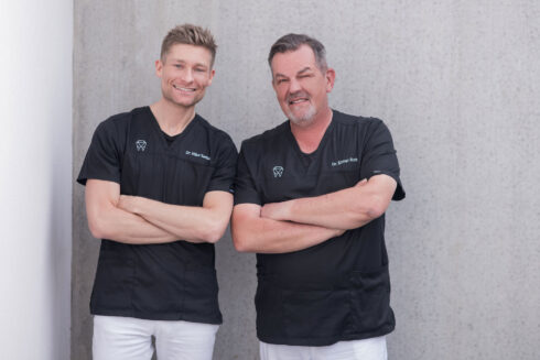 Dr. Mike Siegler und Dr. Elmar Rott stehen lächelnd vor grauer Betonwand mit verschränkten Armen