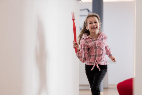 Lachendes Mädchen rennt mit großer, roter Zahnbürste in der Hand durch den Praxisflur