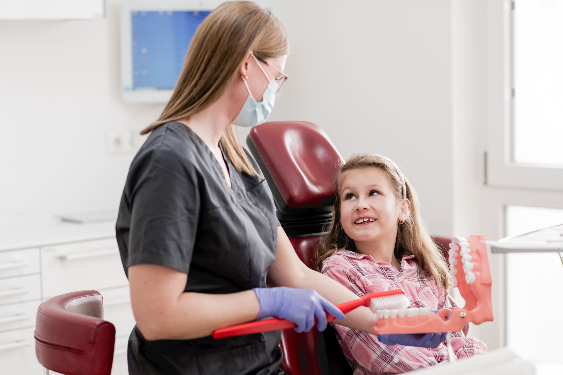 Mädchen liegt auf Zahnarztstuhl. Neben ihr sitzt eine Prophylaxeassistentin mit einer großen roten Zahnbürste